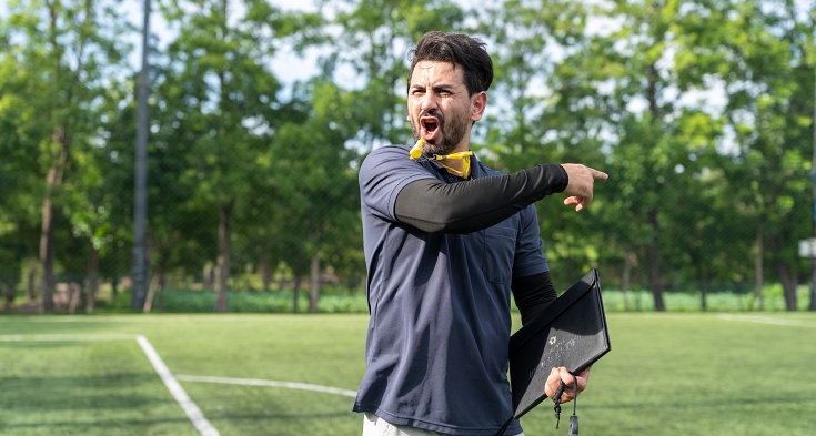 Male sports trainer holding clipboard and whistle yelling instructions inside artificial turf arena, active athletic manager practicing soccer team strategy