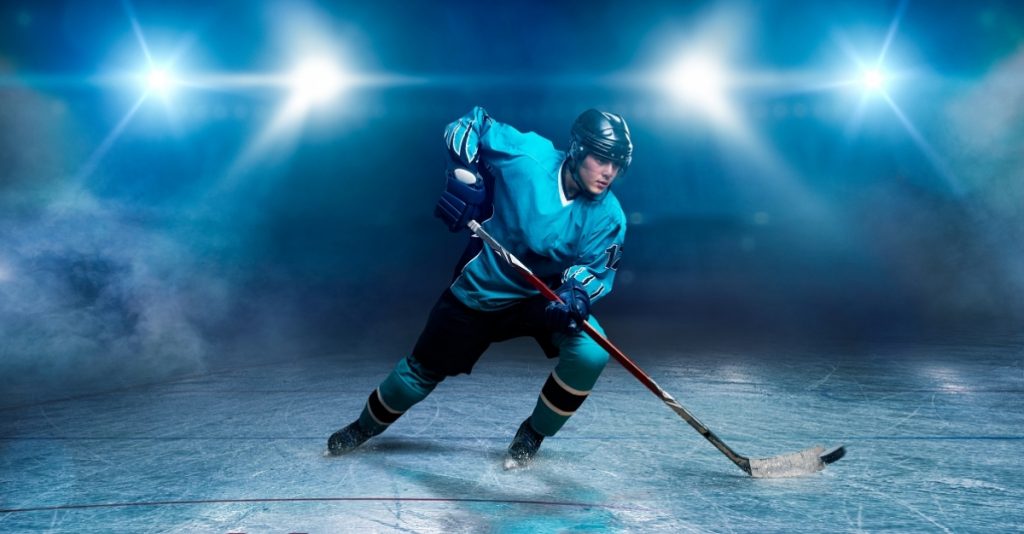 A hockey player on ice, spotlights on background. Male person in helmet, gloves and uniform holds stick in hands