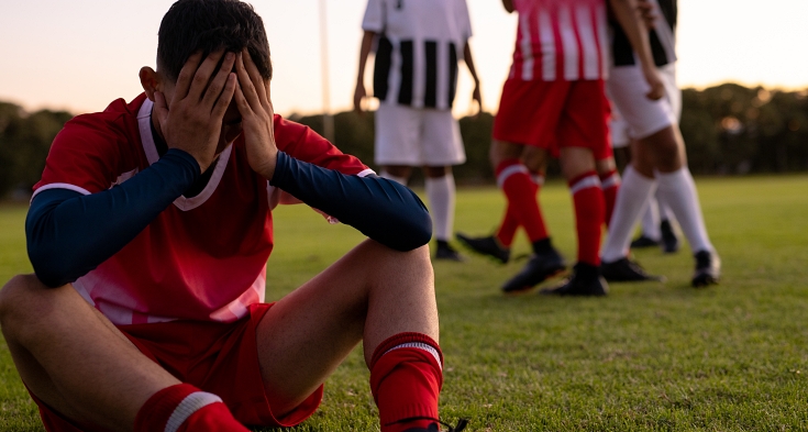 Multiracial sad male soccer player with head in hands sitting on grassy land against clear sky