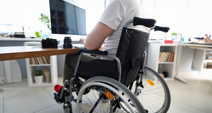 Close-up of man sitting in disabled carriage and looking in computer screen. Adapted workplace and office for people with disabilities. Camera on table. Fulfilling life. Professional specialist concept