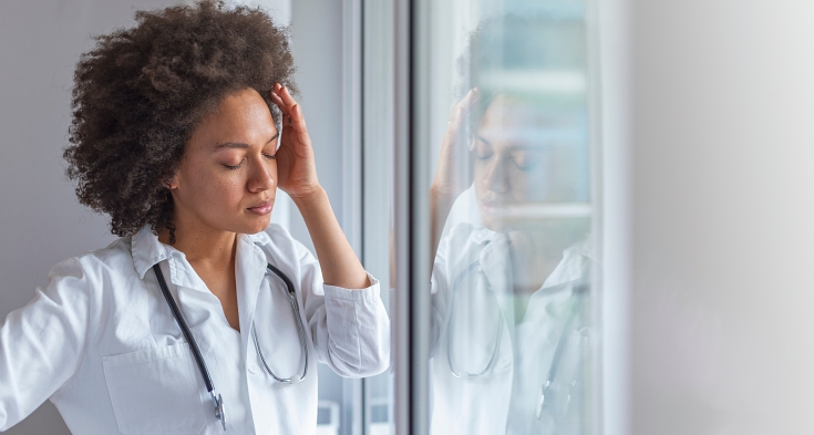 Sad female doctor leaning against the wall in hospital corridor. Female doctor with head pain standing near window. It's a stressful profession. Upset female nurse standing in hospital corridor