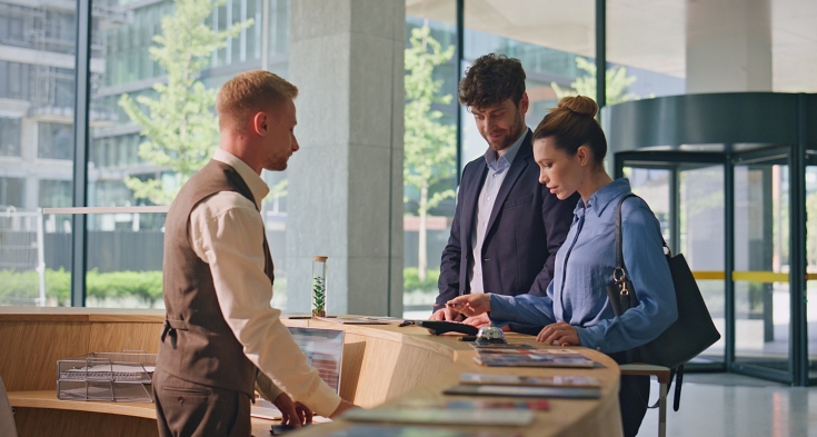 Woman paying at hotel counter with card in modern lobby. Smiling guests checking in settling after arrival. Professional friendly concierge working talking clients at desk. Customer service concept